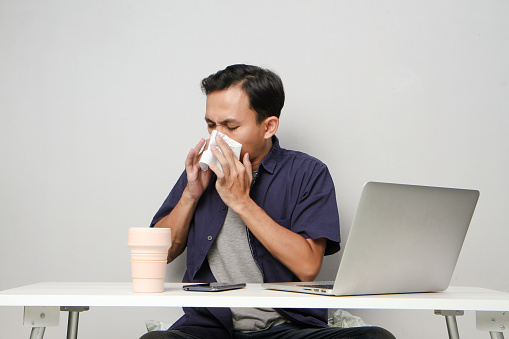 asian man has a runny nose, wipes his nose with tissue paper at workplace while sitting in front of laptop computer. isolated background