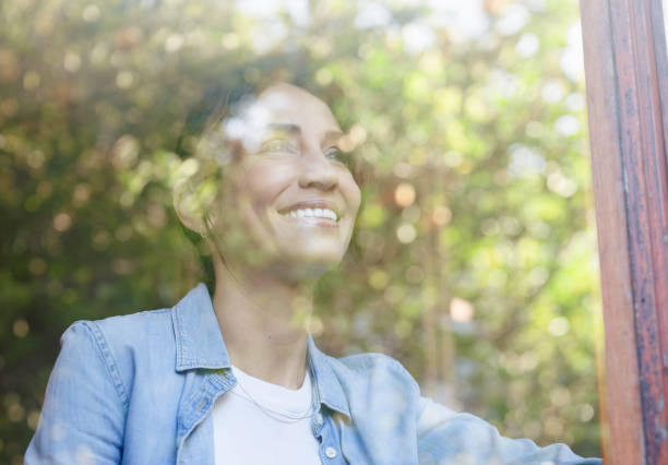 Mature woman relaxing at home and standing next to a window, she is looking away and thinking