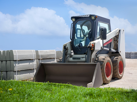 Replacement of the curb stone. Pallets with a curb stones stacked on them and a blue excavator stand on an asphalt road against a blue sky background