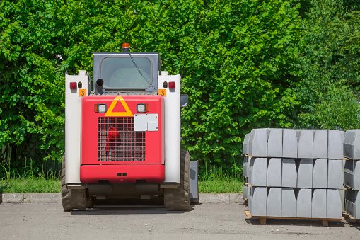 Replacement of the curb stone. Pallets with a curb stone stacked on them and a red and white excavator stand on an asphalt road against a background of dense greenery