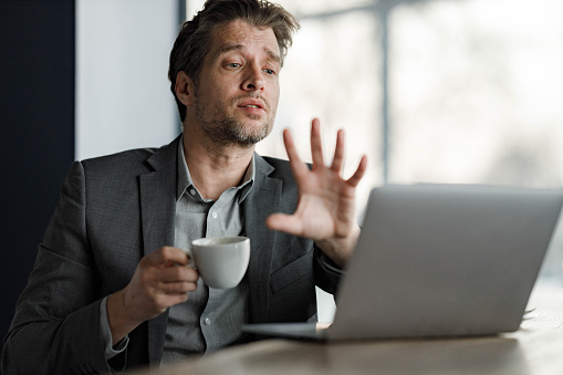 Mid adult businessman drinking coffee while talking to someone during a conference call over a computer in the office. Copy space.