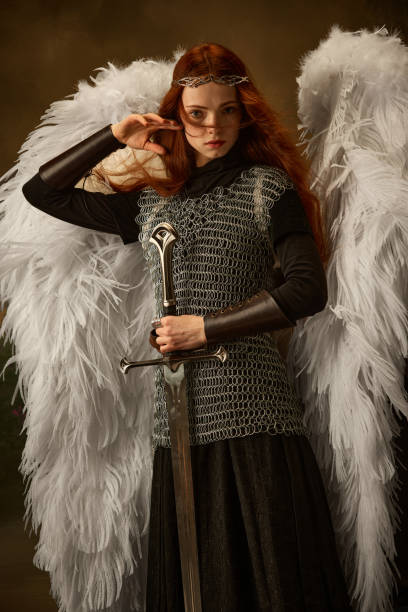 istock Red-haired warrior in chainmail grips sword with white wings looking at camera against vintage studio background. 2149064785