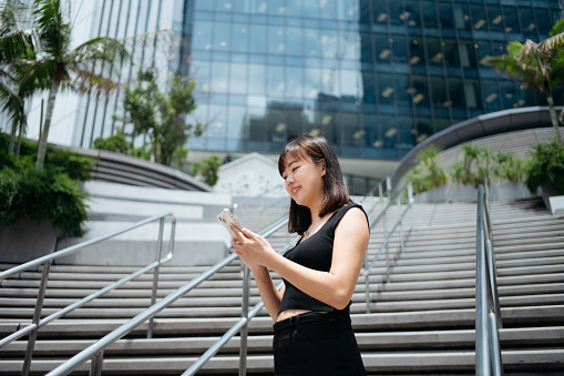 Smiling young Asian businesswoman using smartphone in financial business district. Empowering business with mobile device in the city.