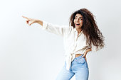 DO YOU SEE THAT. Portrait of amazed pretty curly Latin woman staying near white studio wall background smiling, look at the left and point finger at free place, Copy space for clothes sales