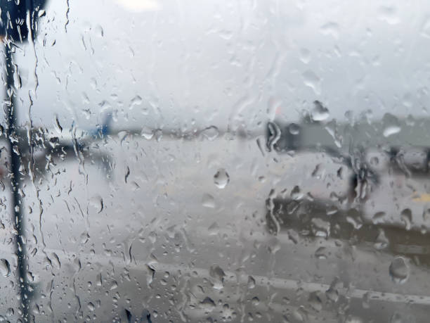 Blurred airplane grounded on the boarding gate, cargo carts around ready for loading before flight in the background, focus on the window with raindrops