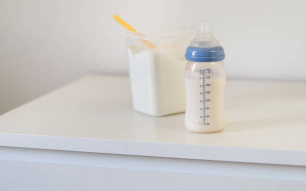 A feeding bottle with baby formula milk on white drawer, dry powder milk in a can with plastic scoop in background