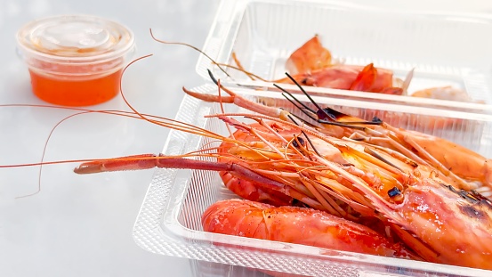 Street food. Plump, grilled prawns in a transparent takeaway box, paired with a tangy dipping sauce, epitomize convenience and flavor, catering to those seeking quick yet indulgent seafood fix
