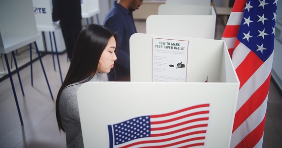 Asian woman comes to voting booth at polling station, makes choice and fills out paper bulletin. US citizen during National Election Day in the United States of America. Civic duty and patriotism.
