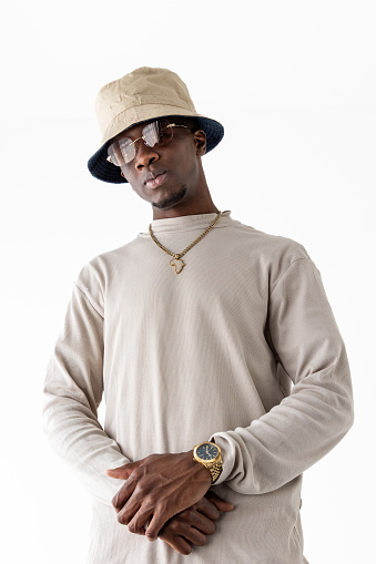 A black man in a studio setting is stylishly dressed in a hat and sunglasses, exuding a cool and confident demeanor.