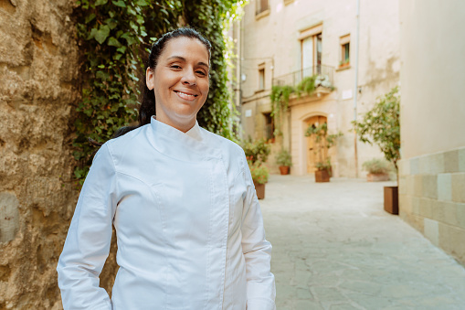 Female chef wears white coat, broadly smiles outdoors on the street. Restaurant worker, culinary gourmet, pastry chef.