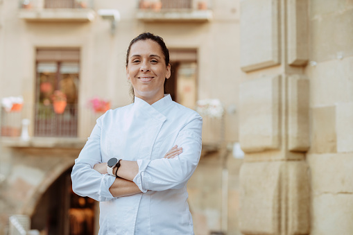 Happy female chef wears white coat, broadly smiles and poses with arms crossed outdoors at city. Restaurant worker, culinary gourmet, pastry chef.