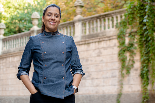 Smiling female chef wears blue coat, poses with hands on pockets outdoors on the street. Restaurant worker, culinary gourmet, pastry chef.