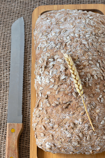 Loaf of bread and wheat on wooden cutting board with knife. Top table view. Close-up. Eating healthy whole grain baked food.