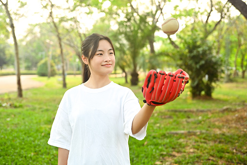 Smiling Asian girl wearing leather glove throwing baseball up into the air.