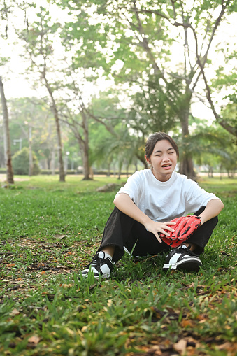 Happy teenage girl wearing leather baseball glove sitting on green lawn during summer day.