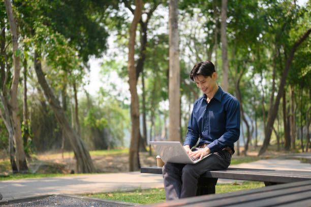 istock Smiling young businessman sitting on bench and using a laptop at public park 2149078968