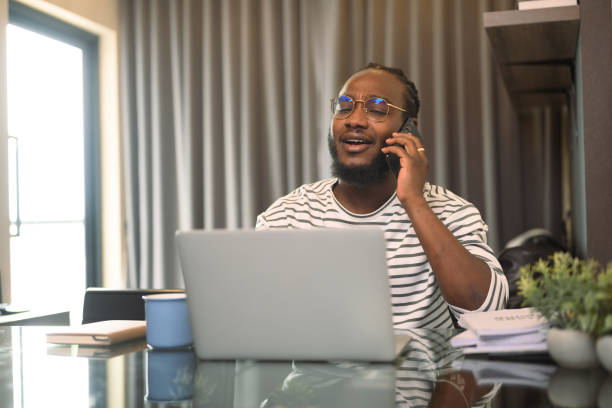 istock Smiling young African man in eyeglasses talking on mobile phone and using laptop at home 2149079012