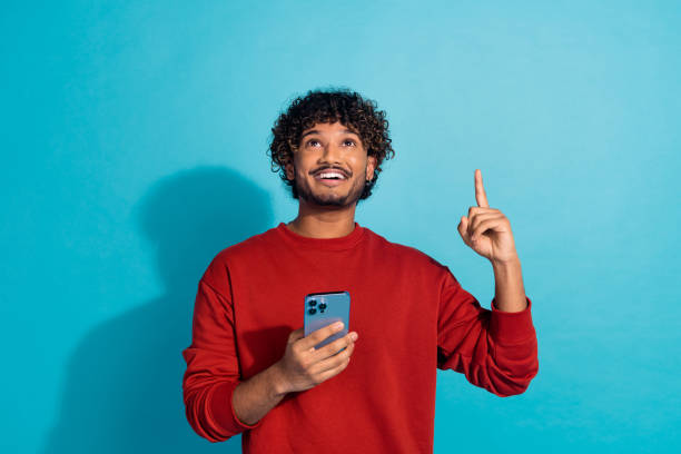 istock Photo of young handsome hispanic guy wearing red pullover directing finger above head use phone recommend media over cyan color background 2149081032