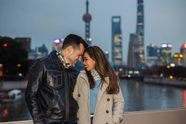 Happy couple  share an intimate moment, their faces close together, against the backdrop of a vibrant cityscape illuminated by the soft glow of evening lights in Shanghai.