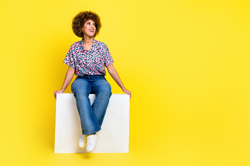 Full length photo of funny dreamy woman wear print shirt sitting cube looking empty space isolated yellow color background.