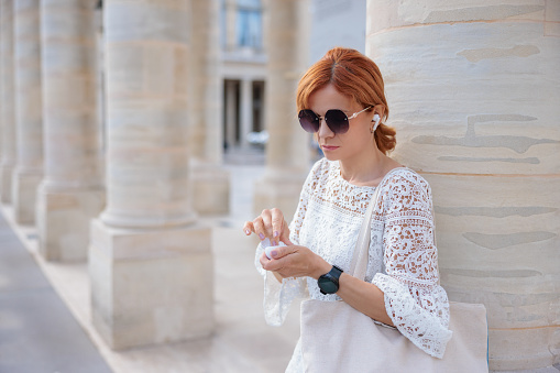 Portrait of beautiful woman standing by architectural column on sightseeing and taking headphones out from a small box, staying connected on the move