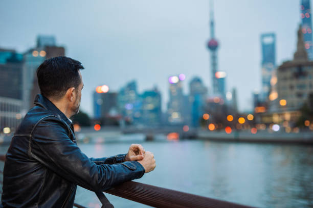 Man clad in a leather jacket leans on a fence, gazing at the illuminated cityscape and tranquil river in Shanghai.