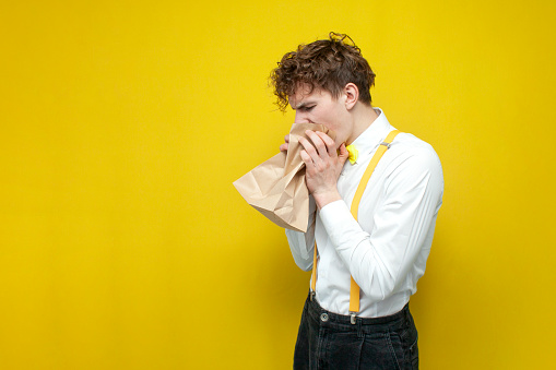 young guy in festive outfit vomits into paper bag on yellow isolated background, the student is ill and sick, intoxication and symptoms