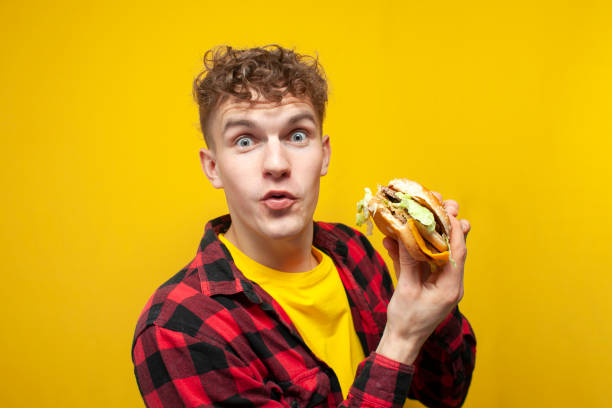 young curly guy student eats big tasty burger on yellow background and shows surprise, satisfied hungry man bites fast food