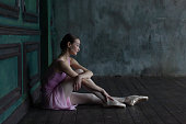 Ballerina in pointe shoes sits thoughtfully on a wooden floor.