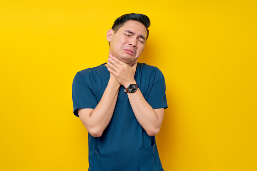 Professional young Asian male doctor or nurse wearing a blue uniform feeling sick and having neck pain, suffering from discomfort after long hours of sedentary isolated on yellow background
