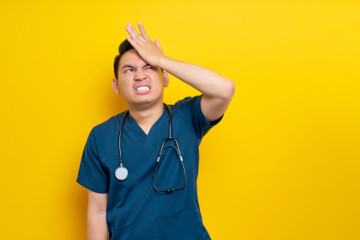 Mistake professional young Asian male doctor or nurse wearing a blue uniform looking aside and hand on his head isolated on yellow background. Healthcare medicine concept