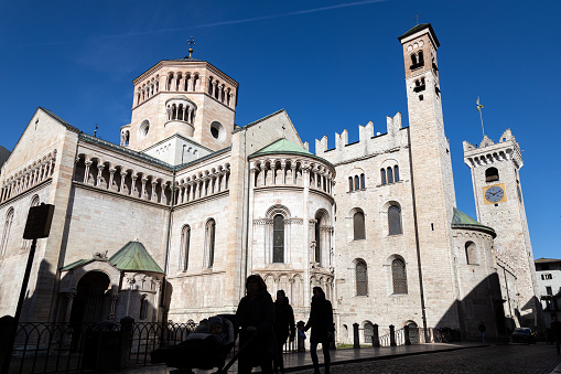 Trento, Trentino-Alto Adige, Italy - Mar 8th, 2024: Silhouette of pedestrians walking by Trento Cathedral in a beautiful sunny day. Clock Tower (Torre Civica or Torre di Piazza) in the background
