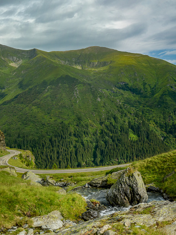 A small mountain stream flowing downhill an alpine pasture through large boulders. The river intersects an asphalt road that winds on the high altitude mountainsides. Fagaras Massif, Carpathia.