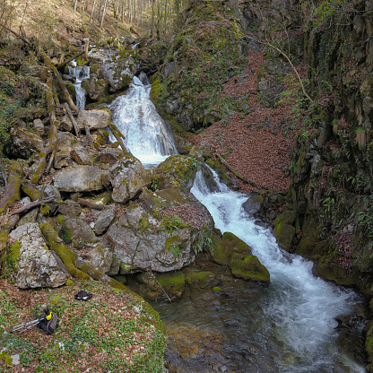 Aerial drone view above a rapid stream flowing through leaves and rocks covered in moss and forming waterfalls. Down on the ground lies photo equipment consisting of a photo camera and a tripod.