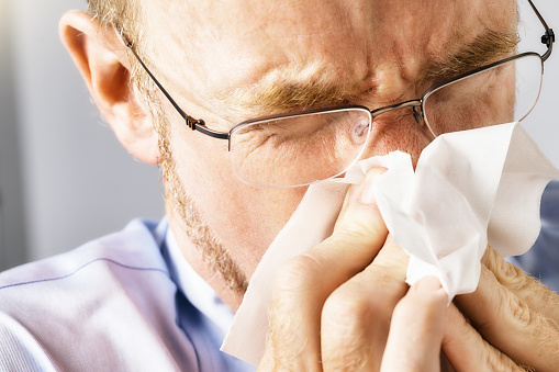 Well-dressed man wearing glasses blows his nose. He has a cold, flu or allergies.