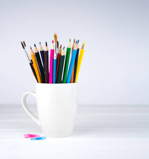 Pencils and brushes in a white mug on a white wooden background. Minimalist composition. Place for text. Selective focus.