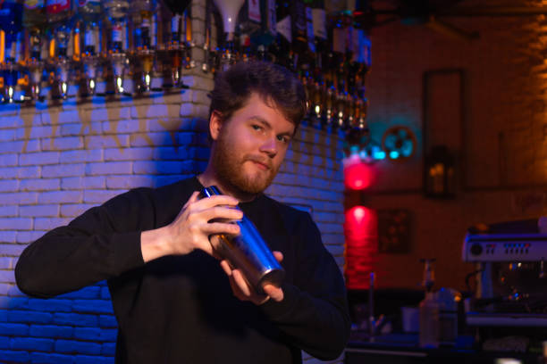 istock Professional bartender with a beard in a black sweatshirt holds a steel shaker behind the bar and looks at the camera. Bartender in a beautiful and atmospheric atmosphere in the bar. 2149092957