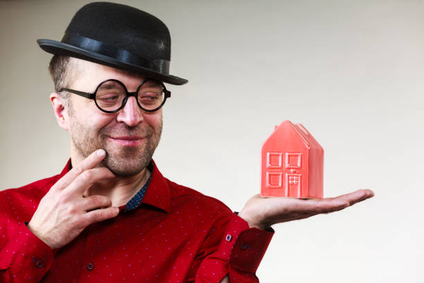 Adult man wearing funny hat and eyeglasses holding small red house model. Guy being real estate agent. Home ownership concept.