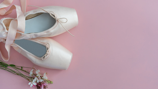 A pair of white ballet shoes are on a pink background. The shoes are tied with a ribbon
