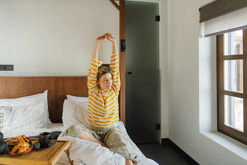 Photo of a young woman waking up, stretching and welcoming morning sun while still in bed, while breakfast platter is by her side