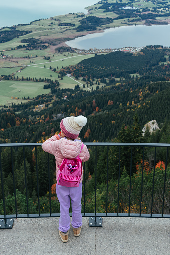 A little girl in a pink jacket and purple pants with a white hat standing on the panoramic view platform in mountains, overlooking lake below, surrounded by dense green forests and rolling hills.