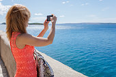 Beautiful woman taking a photo of seascape with smart phone, standing by stone wall on the terrace