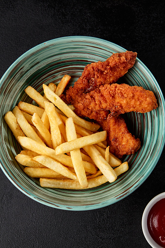 Chicken strips with french fries in bowl on black backdrop. Junk food on dark stone background. Crispy chicken fillet with potatoes in minimal style. Combo with fried chicken and potatoes