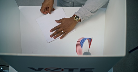 Close up of African American man filling out paper bulletin in voting booth with American flag. Anonymous US citizen votes for president at polling station. Election Day in United States of America.