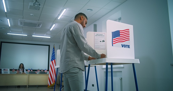 African American male voter walks for registration table at polling station, takes paper ballot. Voting booths with US flag logo and sign calling to vote. Election Day in the United States of America.