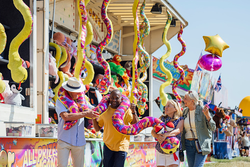 Two senior adult couples visiting an outdoor travelling carnival with different rides and food stalls in the North East of England. They are playing a carnival game together and have won a giant stuffed snake toy as a prize. They are holding the prize together.\n\nVideos are available similar to this scenario.