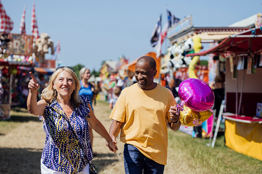 Senior couple visiting an outdoor travelling carnival in the North East of England. They are walking while holding hands. The man is holding balloons and there are rides and various stalls behind them. The woman is pointing ahead of them.\n\nVideos are available similar to this scenario.