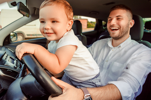 Father playing with toddler son in the car while getting ready for family road trip, boy holding the wheel; family having fun getting ready for travelling to vacation