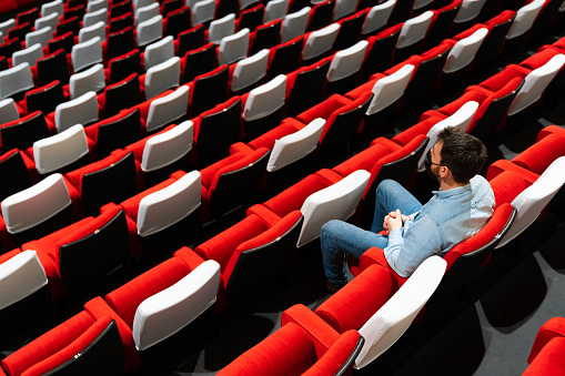 Rear view of a man sitting in an empty theater wearing a protective face mask