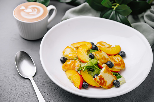 Colorful fruit salad with grilled halloumi, blueberries, and a cup of cappuccino on a dark table